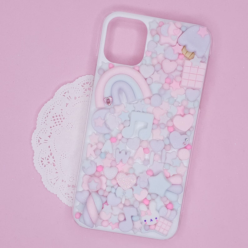 May include: A white phone case decorated with a pink, blue and white pastel rainbow, hearts, stars and music notes. The case is covered in small, colourful sprinkles.