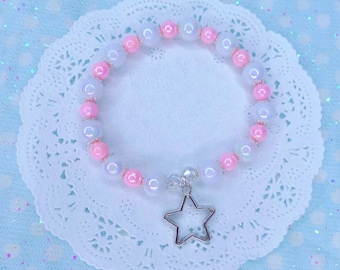 May include: A white and pink beaded bracelet with a silver star charm. The bracelet is on a white doily.
