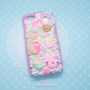 May include: A pink and white phone case with a 3D design featuring a pink cat, a brown bear, a strawberry, a heart, and other pastel-coloured decorations. The case is covered in white frosting and sprinkles.