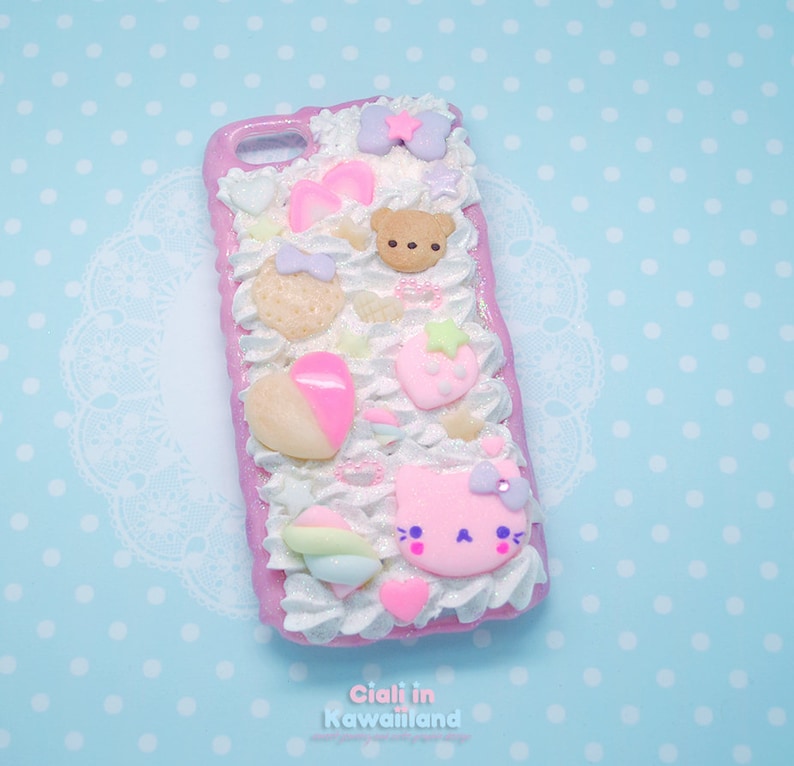 May include: A pink and white phone case with a 3D design featuring a pink cat, a brown bear, a strawberry, a heart, and other pastel-coloured decorations. The case is covered in white frosting and sprinkles.