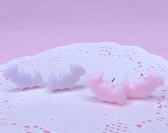 May include: A pair of earrings featuring a bat design. One pair is light purple and the other pair is light pink.