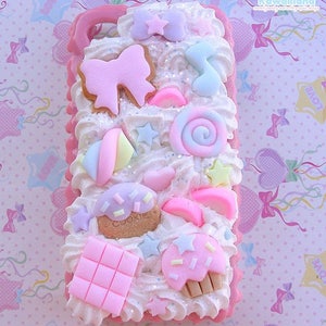 May include: A pink and white phone case decorated with pastel-coloured candy, cookies, and stars. The case has a white background with pink accents and a white border. The text "Ciati in Kawaii land" is printed in the top right corner.