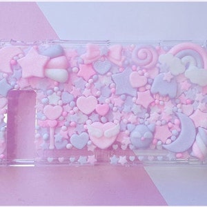 Puede incluir: Una funda de pl&aacute;stico transparente decorada con formas 3D en rosa pastel, morado y blanco, incluyendo estrellas, corazones, nubes y una luna creciente. La funda est&aacute; dise&ntilde;ada para contener una consola Nintendo Switch.