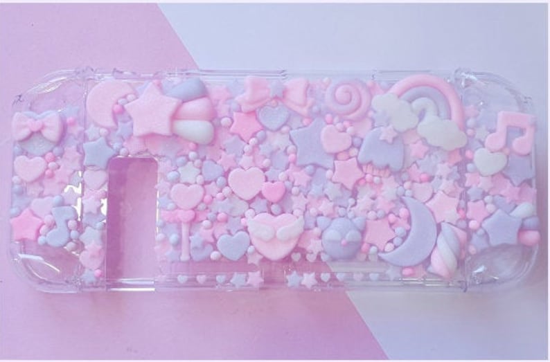 Puede incluir: Una funda de pl&aacute;stico transparente decorada con formas 3D en rosa pastel, morado y blanco, incluyendo estrellas, corazones, nubes y una luna creciente. La funda est&aacute; dise&ntilde;ada para contener una consola Nintendo Switch.