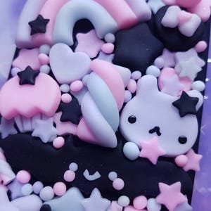 Op de afbeelding: Een close-up van een zwarte telefoonhoes die is versierd met pastelroze, paarse en blauwe 3D-versieringen. De versieringen omvatten een regenboog, sterren, harten, een konijn en een vleermuis.
