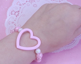 May include: A pink heart-shaped charm bracelet with white and pink beads. The bracelet is on a pink background.
