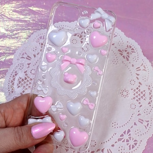 Könnte beinhalten: Eine transparente Handyhülle mit einem rosa und weißen Design. Die Hülle ist mit Herzen, Schleifen und Spitze verziert.