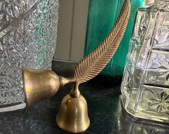May include: A gold-colored bell with a leaf-shaped handle. The bell is sitting on a black surface next to a clear glass bottle and a green glass bottle.