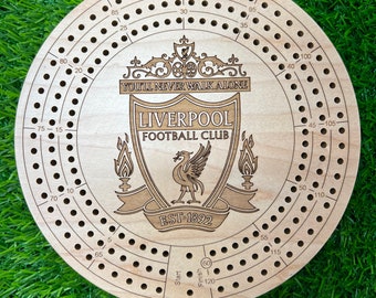 May include: A wooden circular game board with a Liverpool Football Club crest in the center. The crest features a red bird with a crown, a shield with the words "You'll Never Walk Alone" and "Liverpool Football Club", and the text "EST. 1892". The board has a series of numbered holes around the edge, starting at 5 and going up to 120.