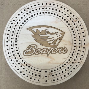 May include: A round wooden cribbage board with a laser-etched beaver logo and the word "Beavers". The board has numbered holes for scoring, with a start and finish point. The board measures approximately 12 inches in diameter.