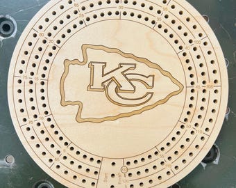May include: Circular wooden cribbage board with a laser-cut Kansas City Chiefs logo in the center. The board features numerous holes for pegs and is marked with numbers for scoring. The board is made of light-colored wood.