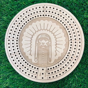 May include: A wooden cribbage board with a Native American headdress design. The board has a circular shape and features a numbered track for scoring points. The numbers on the board range from 5 to 120. The board also includes a start and finish point.