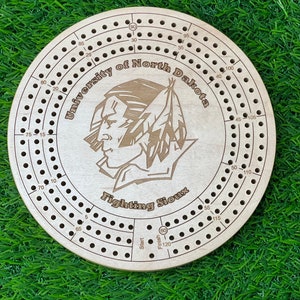 May include: A wooden cribbage board with a University of North Dakota logo and the words "Fighting Sioux". The board has a circular shape and is made of light brown wood. The logo is a Native American head with a feather headdress.