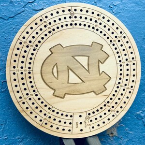 May include: A wooden cribbage board with a circular design featuring the University of North Carolina Tar Heels logo. The board has a numbered track with holes for pegs and the numbers 1-120.