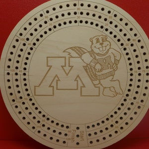 May include: Circular wooden cribbage board with a laser-etched design. The board features a cartoon beaver wearing a sweater with the letter "M" and the University of Minnesota logo. The board has multiple rows of holes for pegging and the words "Start" and "Finish".