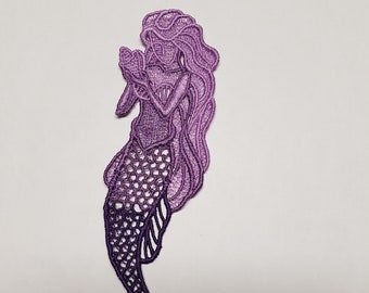 May include: A purple embroidered mermaid patch. The mermaid has long flowing hair and is holding a shell. The mermaid's tail is a darker shade of purple, with a detailed scale pattern. The patch is designed to be sewn or ironed onto fabric.