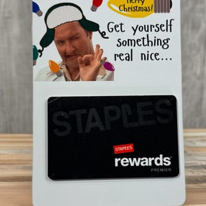 May include: A white gift card with a black Staples Rewards card. The top of the card has a humorous image of a man wearing a hat with Christmas lights and the text "Merry Christmas! Get yourself something real nice..."