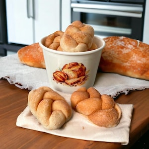 May include: A white ceramic bowl with a bread design on the side, filled with braided bread rolls. Two additional braided bread rolls are on a white linen napkin next to the bowl. A loaf of bread is on the table behind the bowl.