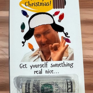 May include: A rectangular card featuring a printed image of a person wearing a black and white trapper hat, making an okay hand gesture, surrounded by colorful Christmas lights. Text reads Merry Christmas and Get yourself something real nice.