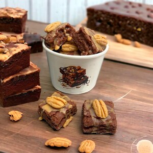 May include: A close-up of a white paper cup filled with chocolate brownies topped with pecans and caramel sauce. The brownies are sitting on a wooden surface with other brownies and pecans.
