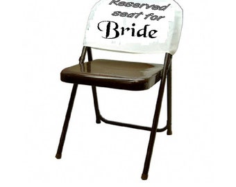 Puede incluir: Una silla plegable de metal marrón con un respaldo de tela blanco. El respaldo tiene el texto "Asiento reservado para la novia" impreso en él.
