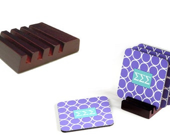 Puede incluir: Conjunto de cuatro posavasos de color morado y blanco con un patrón de círculos y las letras "ΣΣΣ" en color turquesa. Los posavasos se muestran en un soporte de madera.