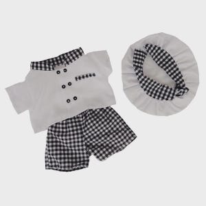May include: A white chef's outfit for a child, including a short-sleeved jacket with black buttons, matching shorts, and a chef's hat. The jacket has a black and white checkered collar and pocket trim. The shorts and hat trim feature the same checkered pattern.