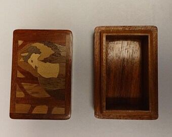 May include: A rectangular wooden box with a lid, featuring a marquetry design of a figure. The box is open, revealing the interior. The wood has a warm, reddish-brown hue, with lighter wood accents in the design.