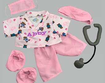 Puede incluir: Disfraz de doctor de oso de peluche rosa con una bata de quirófano, pantalones, gorro, máscara y estetoscopio. La bata de quirófano tiene un fondo rosa con pequeños corazones y osos de peluche. El nombre Abby está bordado en la bata de quirófano en rosa.