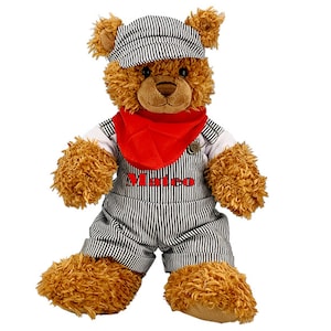 Peut inclure: Un ours en peluche brun portant une salopette rayée noire et blanche, un bandana rouge et une casquette rayée noire et blanche. La salopette a le nom "Mateo" brodé sur le devant.