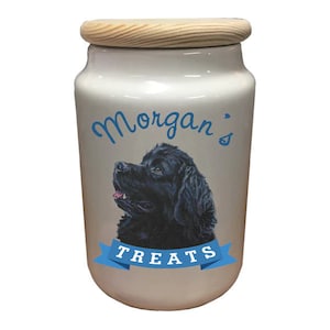 May include: White ceramic dog treat jar with a wooden lid. The jar has a black and white illustration of a Newfoundland dog with the text "Morgan's Treats" on a blue ribbon.