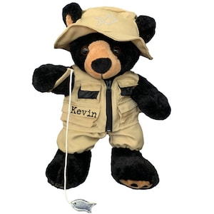 May include: Black plush bear wearing a tan fishing vest and hat with a fish design. The bear is holding a fishing pole with a fish at the end. The vest has a zipper and pockets. The bear is personalized with the name "Kevin".