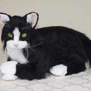 May include: A plush, lifelike cat figurine in a relaxed pose. The cat has black fur with white markings on its face, paws, and tail. It has green eyes and long white whiskers. The cat is lying down on a patterned surface.