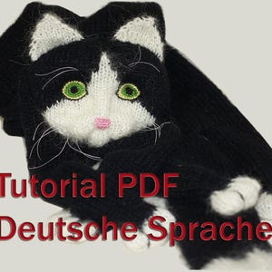 May include: Black and white knitted cat scarf with green eyes and a pink nose. The scarf has the text "Tutorial PDF Deutsche Sprache" on it.
