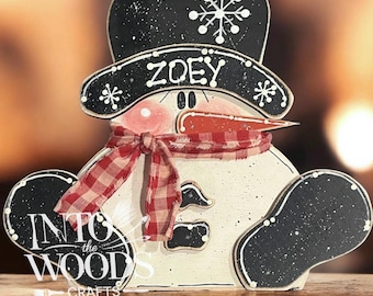 May include: A wooden snowman decoration featuring a black top hat with a white snowflake, the name ZOEY in white lettering, a red and white checkered fabric scarf, a carrot-shaped nose, and black arms, with the text INTO THE WOODS CRAFTS.