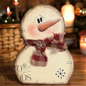 May include: A wooden snowman cutout with a cream-colored finish, featuring a painted orange carrot nose, black eyes, and rosy cheeks. A frayed red and tan plaid fabric scarf is tied around the neck. A black snowflake design is on the lower body.