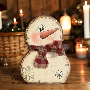 May include: A wooden snowman decoration with a distressed, off-white finish. It features a carrot nose, black eyes, and a red and brown plaid scarf. The base has a black snowflake and the text "Into Woods Crafts".