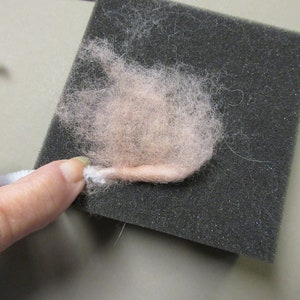 May include: A close-up of a pink wool fiber being felted on a black foam mat. The fiber is being rolled and compressed to create a felt ball.