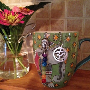 May include: A green ceramic mug with an elephant design and a silver charm with the Om symbol hanging from it. The mug has a teal handle and is sitting on a wooden surface.