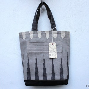 May include: A gray and black tote bag with a black leather bottom and a white tag hanging from the handle. The bag has a geometric pattern of black triangles on a gray background.