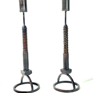 以下が含まれることがあります： ユニークなデザインの黒い金属製のキャンドルホルダー2つ。各ホルダーは、湾曲したアーチが付いた円形のベース、正方形のナット、らせん状のデザインの長い薄い金属棒を備えています。各棒の上部には円筒形のキャップが付いています。