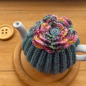 May include: A blue knitted teapot cosy with a colourful crocheted flower on top. The flower has pink, yellow and green stripes and a blue button in the centre.
