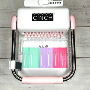 May include: A white and pink Cinch binding machine with a ruler and five different colored binding strips. The strips are labeled "AS PORTRAIT", "AS LANDSCAPE", "AS PORTRAIT", "AS LANDSCAPE", and "26 LANDSCAPE".