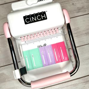 May include: White and pink Cinch binding machine with a silver handle. The machine has a ruler in inches and five different coloured binding spines.