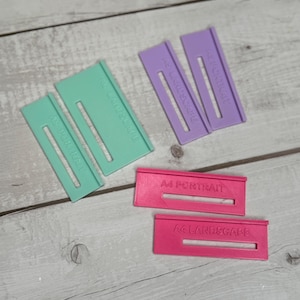May include: Three sets of paper size guides for cutting paper. The guides are made of plastic and are in different colours: green, purple, and pink. The guides are labelled with the paper size and orientation: A4 Portrait, A4 Landscape, A3 Portrait, A3 Landscape.