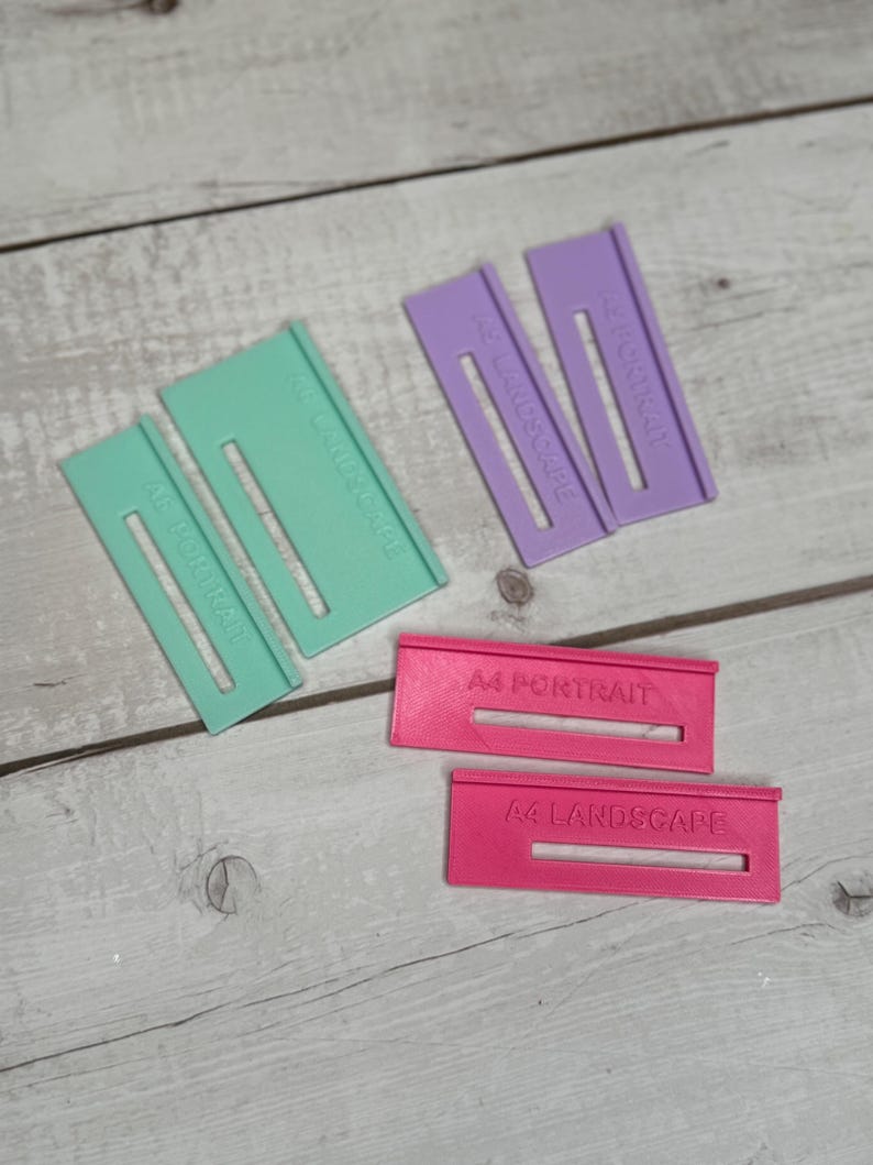 May include: Three sets of paper size guides for cutting paper. The guides are made of plastic and are in different colours: green, purple, and pink. The guides are labelled with the paper size and orientation: A4 Portrait, A4 Landscape, A3 Portrait, A3 Landscape.
