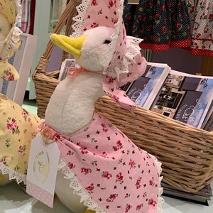 May include: A white plush goose wearing a pink and white floral bonnet and a pink floral apron with white lace trim. The goose is sitting in a wicker basket with a tag that says "Cotton Tails".