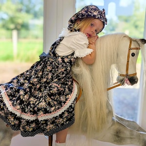 May include: A young person wearing a floral print dress and matching bonnet, resting on a white rocking horse. The dress features a black background with pink and white floral patterns, and a white lace trim.