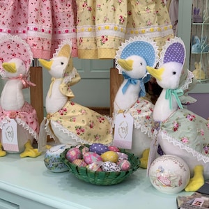 Può includere: Quattro statuette di oche di peluche con cappellini e vestiti floreali. Le oche hanno becchi e zampe gialle, e ognuna indossa un cappellino e un vestito di colore diverso. Un cestino verde di uova di Pasqua decorate è davanti alle oche.