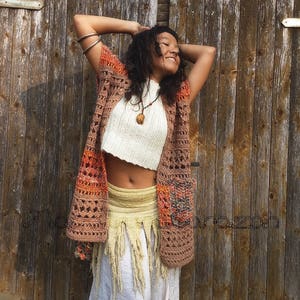 Puede incluir: Una mujer lleva un chaleco de crochet marrón y naranja con un top de crochet blanco y una falda larga blanca con flecos. Está de pie frente a una pared de madera.